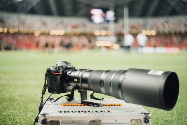 A camera with a really long zoom lens sitting on a silver case, on the field in a stadium